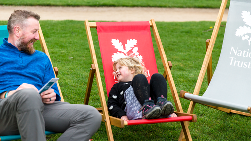 A man on a blue deckchair in blue jumper, with his daughter on a red deckchair, grinning at him